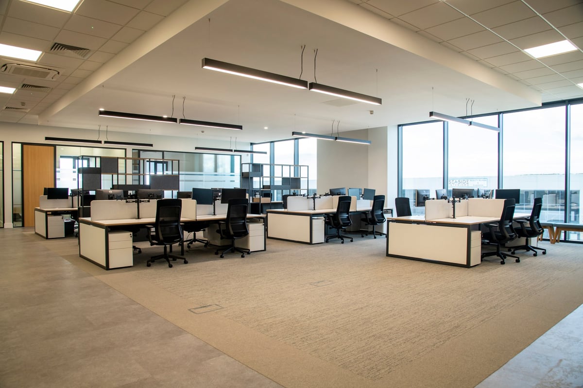 Modern open-plan office space with light wood desks, black chairs, pendant lighting, and floor-to-ceiling windows overlooking a city skyline