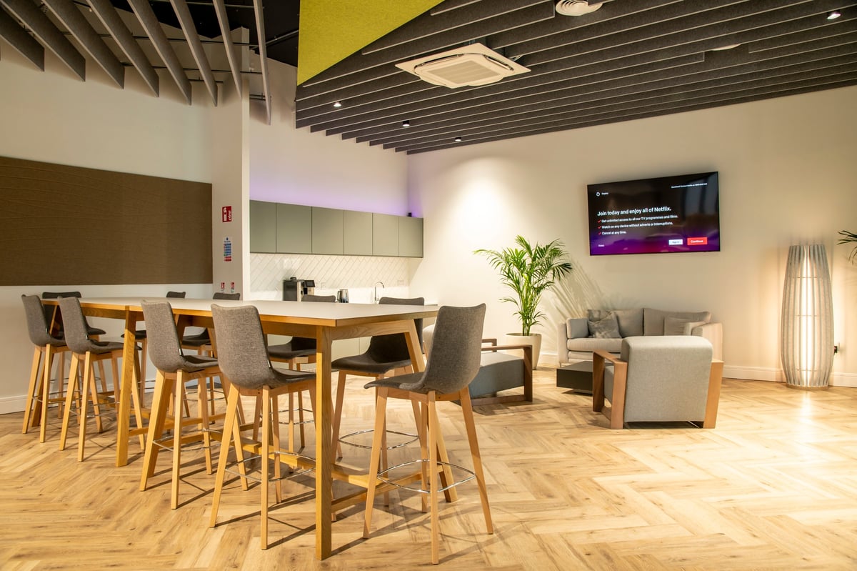 Modern office break room with wooden bar seating, gray upholstered chairs, lounge area with sofa, kitchenette, potted plant, and digital display screen