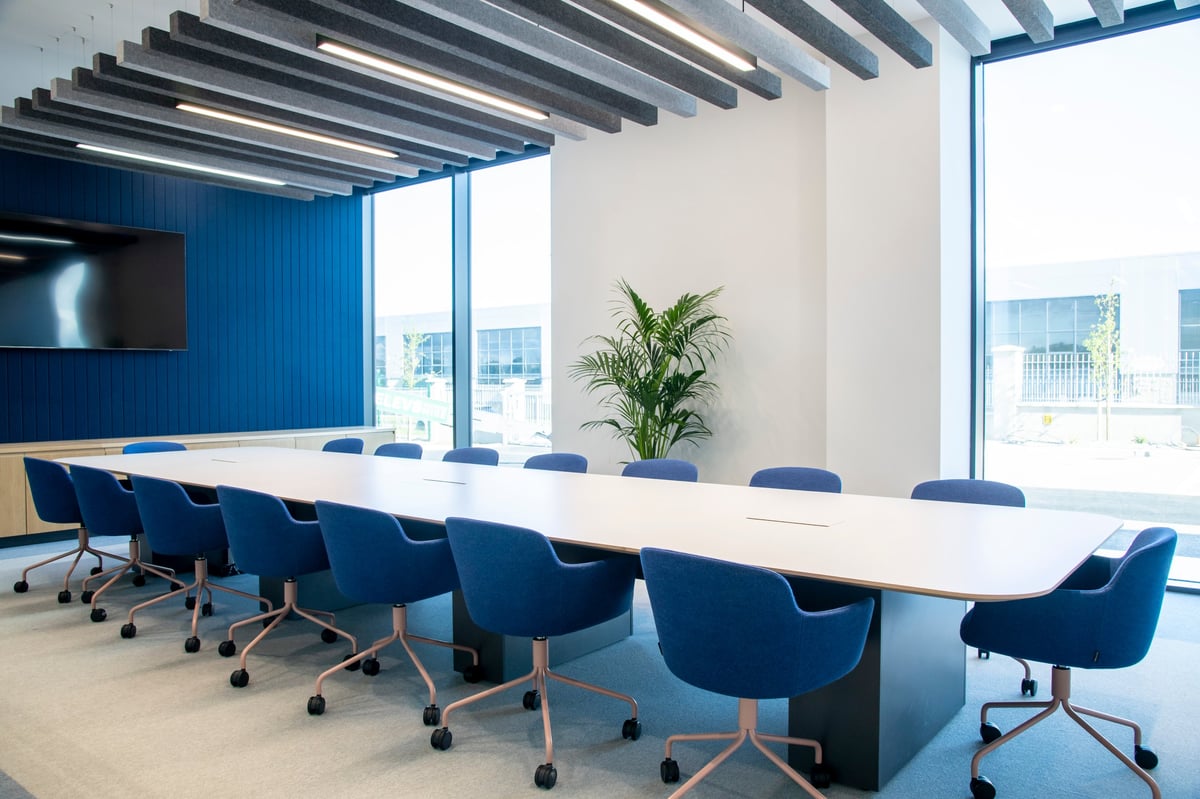 Modern conference room with long white table, blue chairs, exposed ceiling beams, and floor-to-ceiling windows overlooking a city skyline