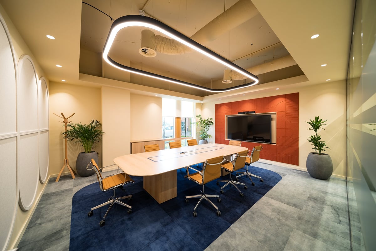 Modern office conference room with large white table, wood chairs, red accent wall with TV, and architectural lighting fixtures
