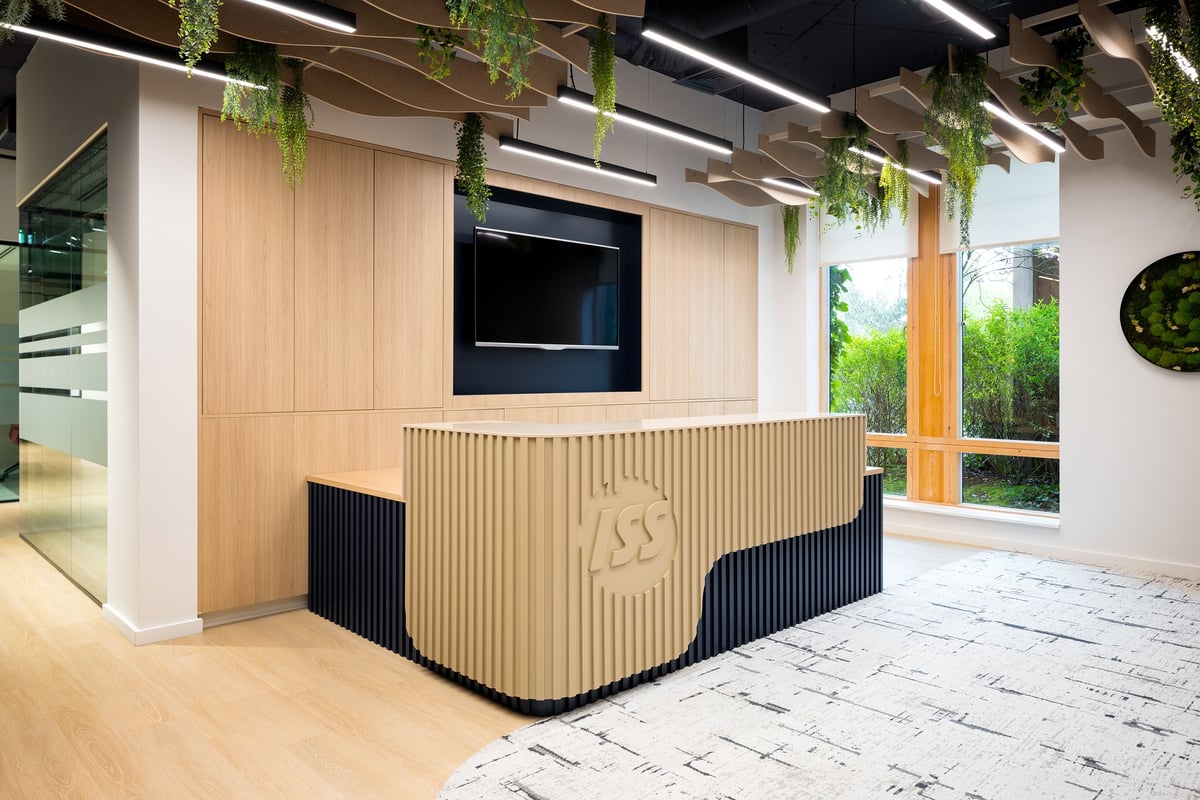 Modern office reception desk with wood and black paneling, mounted TV screen, hanging plants, and large windows in bright minimalist space