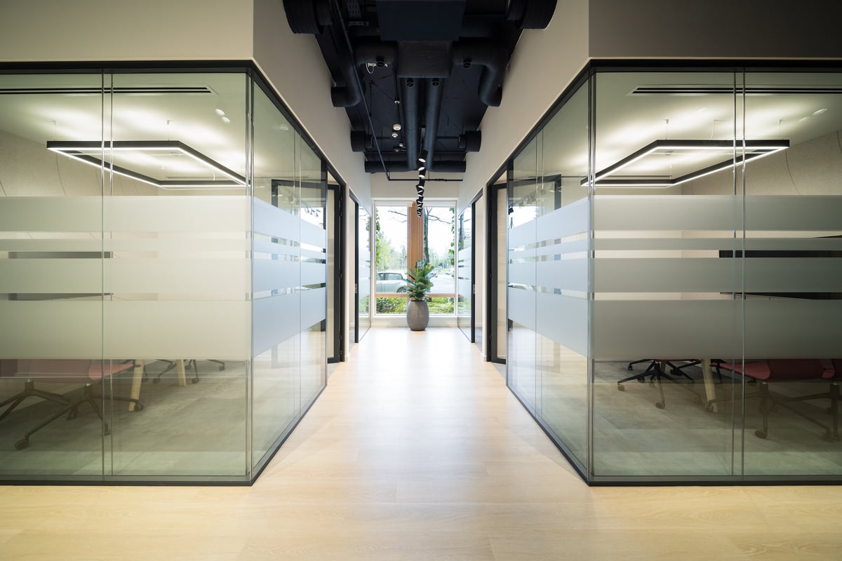 Modern office corridor with glass-walled meeting rooms on both sides and a central walkway leading to a green outdoor area