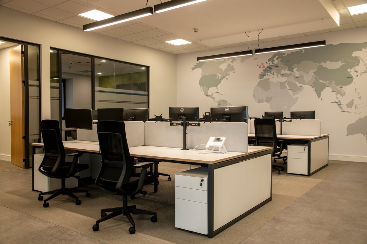 Modern office space with desks, black ergonomic chairs, computer monitors, and a world map on the wall under fluorescent lighting